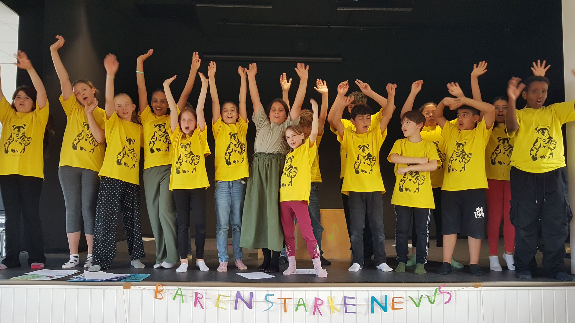 Eine Gruppe von Kindern steht mit erhobenen Armen auf einer Bühne. Die Kinder tragen passende gelbe Shirts mit schwarzen Motiven. Die Bühnenfront ziert ein Schild mit der Aufschrift „BÄRENSTARKE NEWS“.